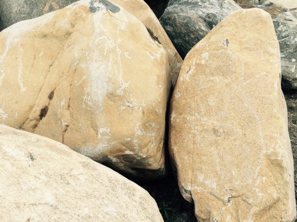 A pickaxe with a yellow handle leaning against a pile of large rocks on a dirt surface, illustrating construction or landscaping activity in Long Island, NY.