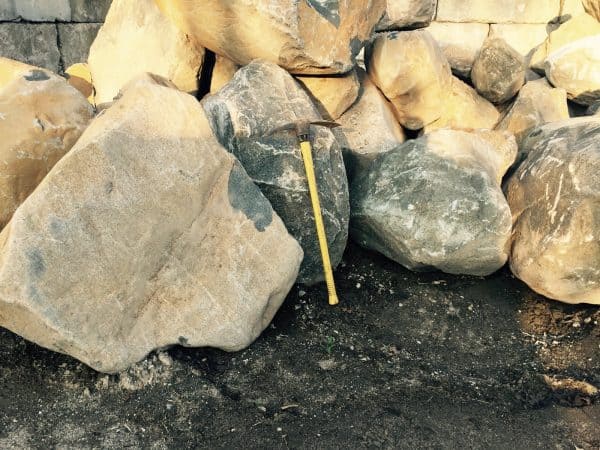 A pickaxe with a yellow handle leaning against a pile of large rocks on a dirt surface, illustrating construction or landscaping activity in Long Island, NY.