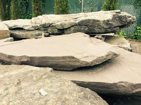 Stacked flat stone slabs with rough edges in an outdoor yard near a green chain-link fence, used for landscaping or construction in Long Island, NY.