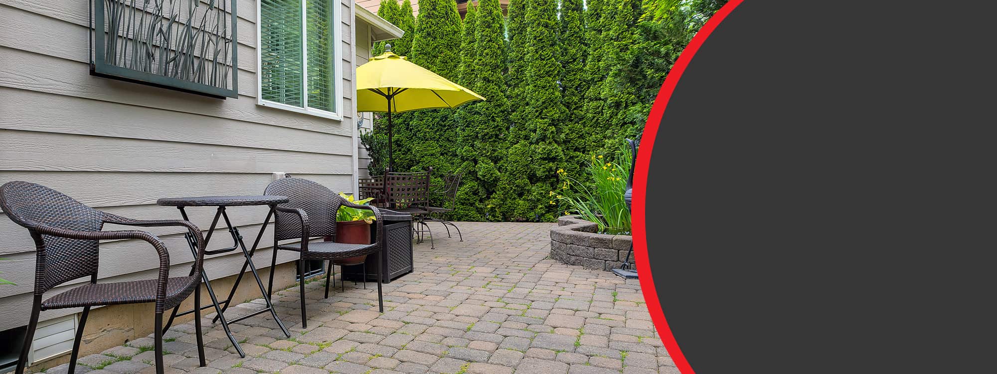 A stone patio with wicker chairs, a yellow umbrella, and lush green hedges providing privacy in a residential setting in Long Island, NY.