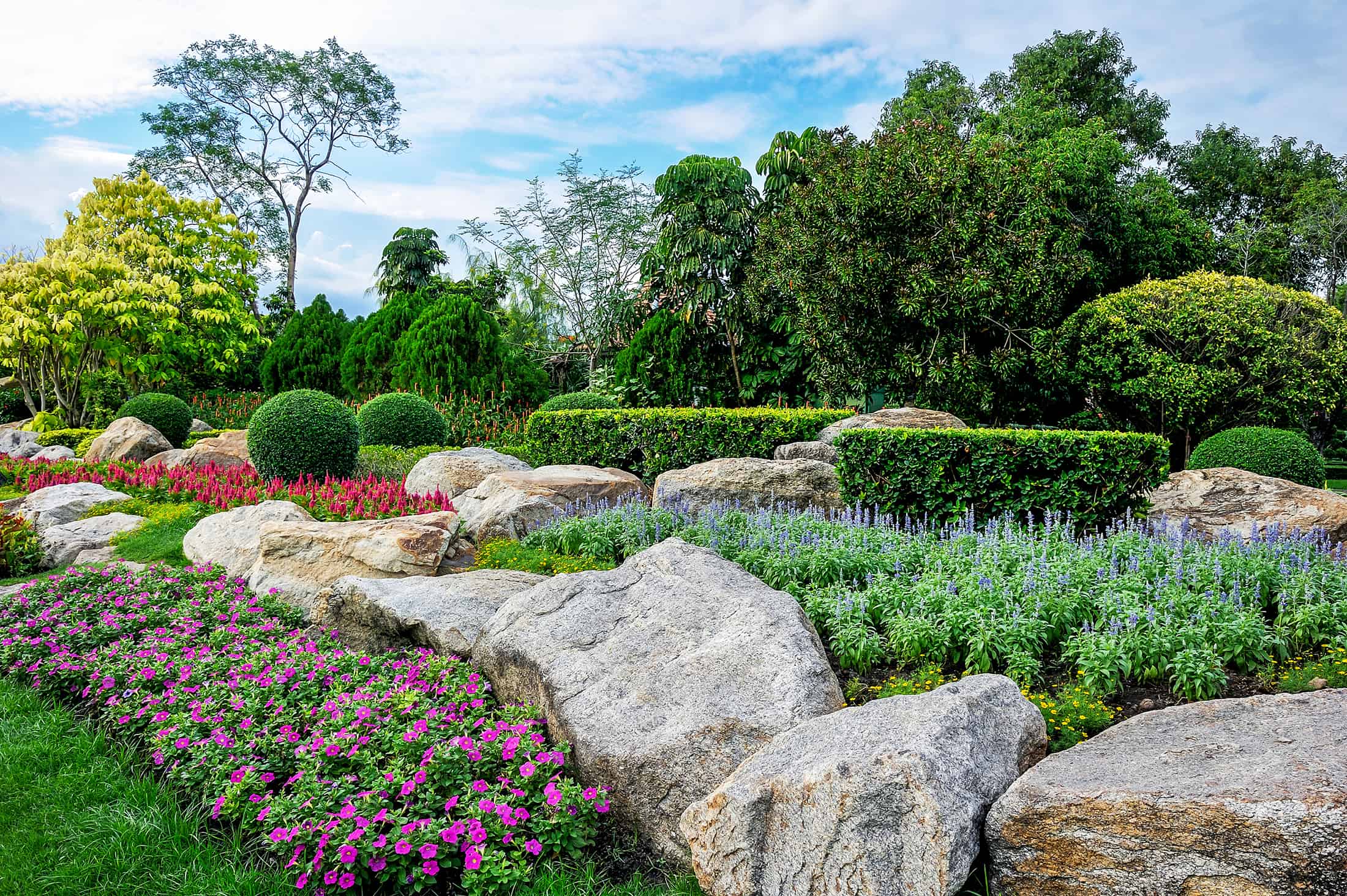 Beautifully landscaped garden with vibrant flowers, large rocks, and tall trees under a partly cloudy sky in Long Island, NY.