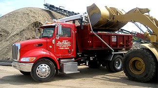 A red dump truck labeled 'John Tuttle Trucking & Excavating' being loaded with dirt by a yellow front loader at a construction site in Long Island, NY.