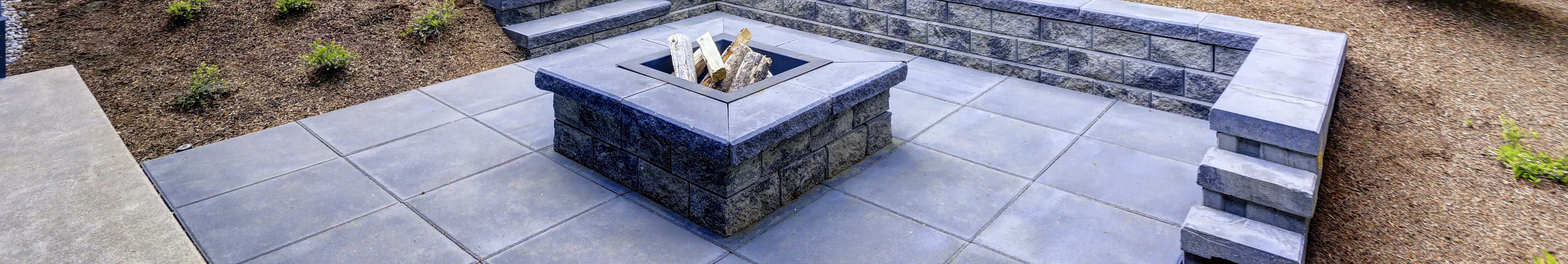 A stylish outdoor patio in Long Island, NY featuring a central stone fire pit, raised seating walls, and landscaped surroundings with mulch and greenery.