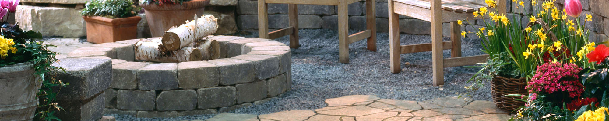 A circular stone fire pit surrounded by a cushioned curved bench in a natural garden setting in Long Island, NY, creating a cozy outdoor gathering space.